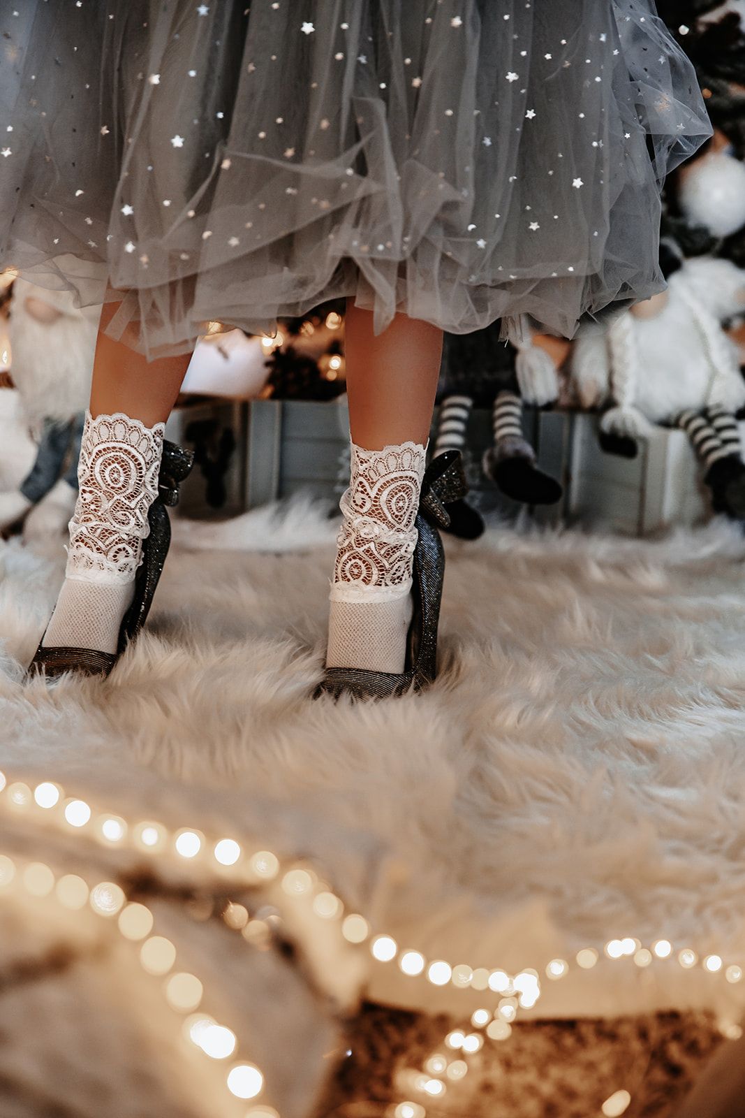 Child in gray tulle dress wearing white lace socks and black shoes standing on fluffy white rug with Christmas lights.
