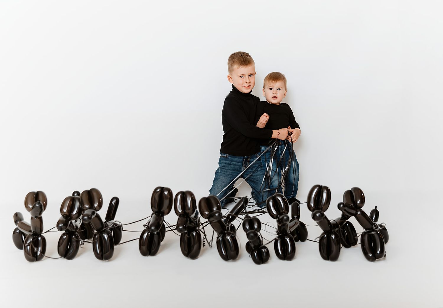 Two young brothers in matching black outfits stand surrounded by black balloon dogs on white background.