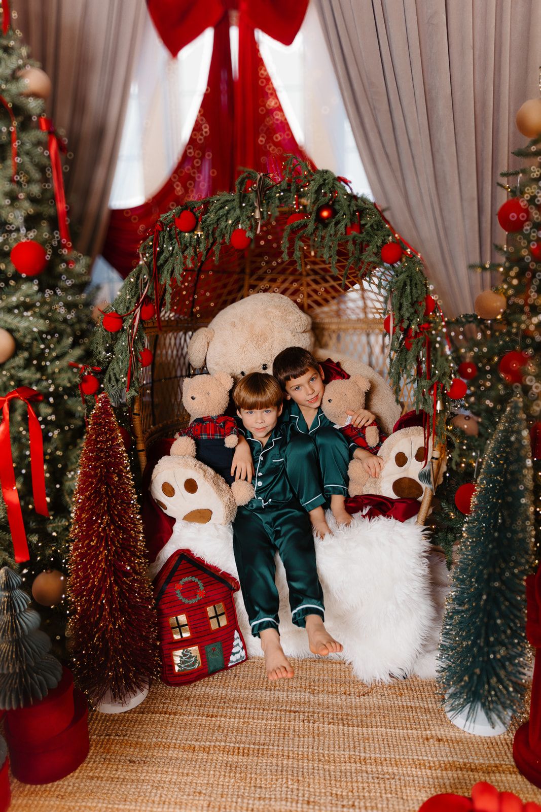 Two children in green satin pajamas cuddle with teddy bears in a festive Christmas chair surrounded by decorated trees.