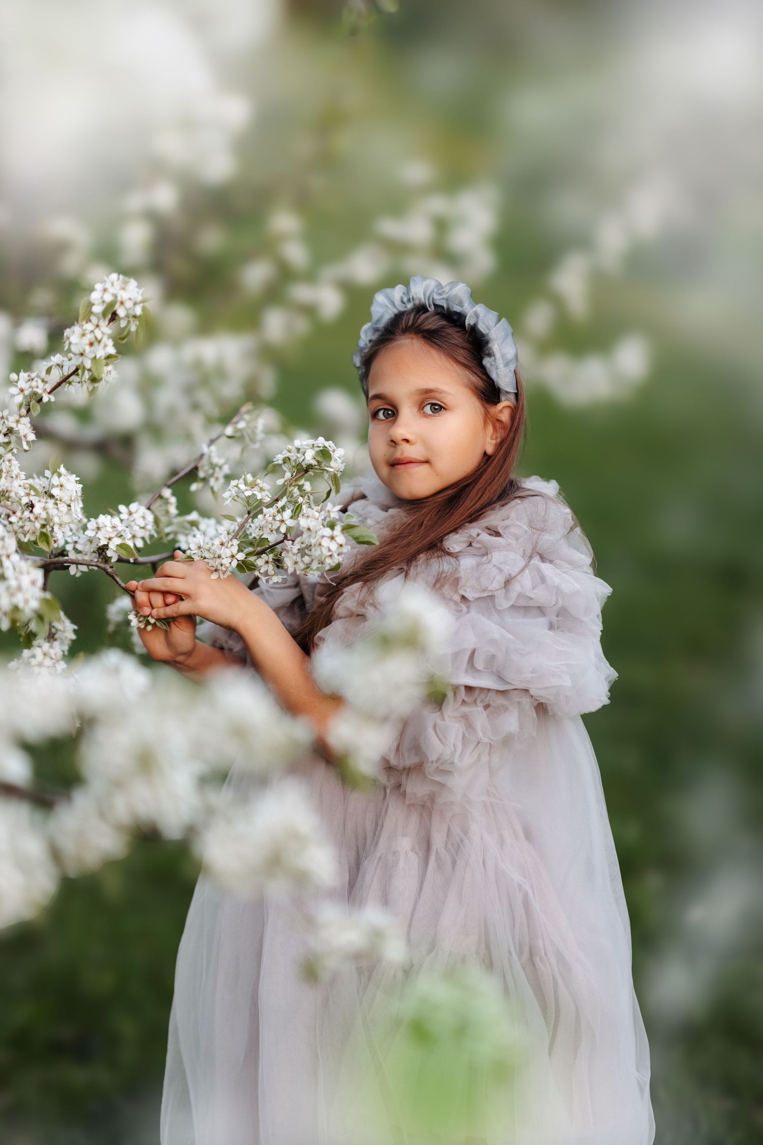 A young girl in a white tulle dress and floral crown poses gracefully among blooming white spring tree branches.