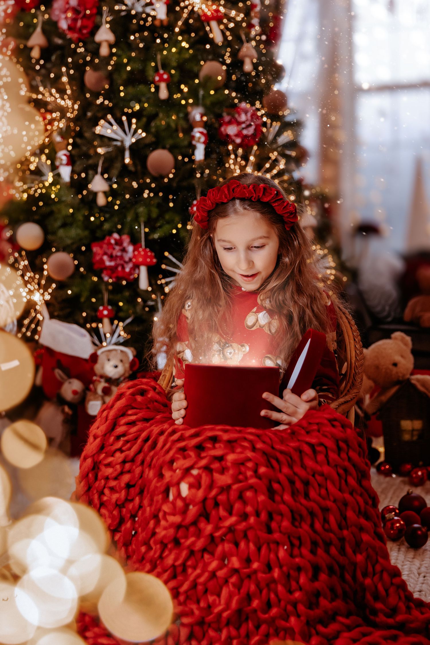 A girl with a red headband opens a glowing gift box while seated near a beautifully decorated Christmas tree.
