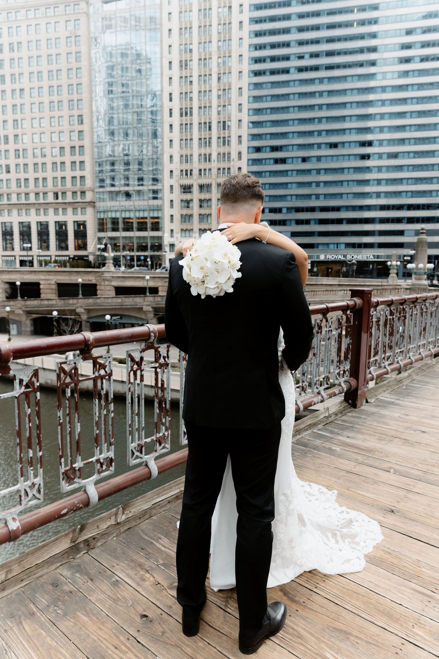 Bride and groom embrace from behind on Chicago Riverwalk with white floral bouquet.