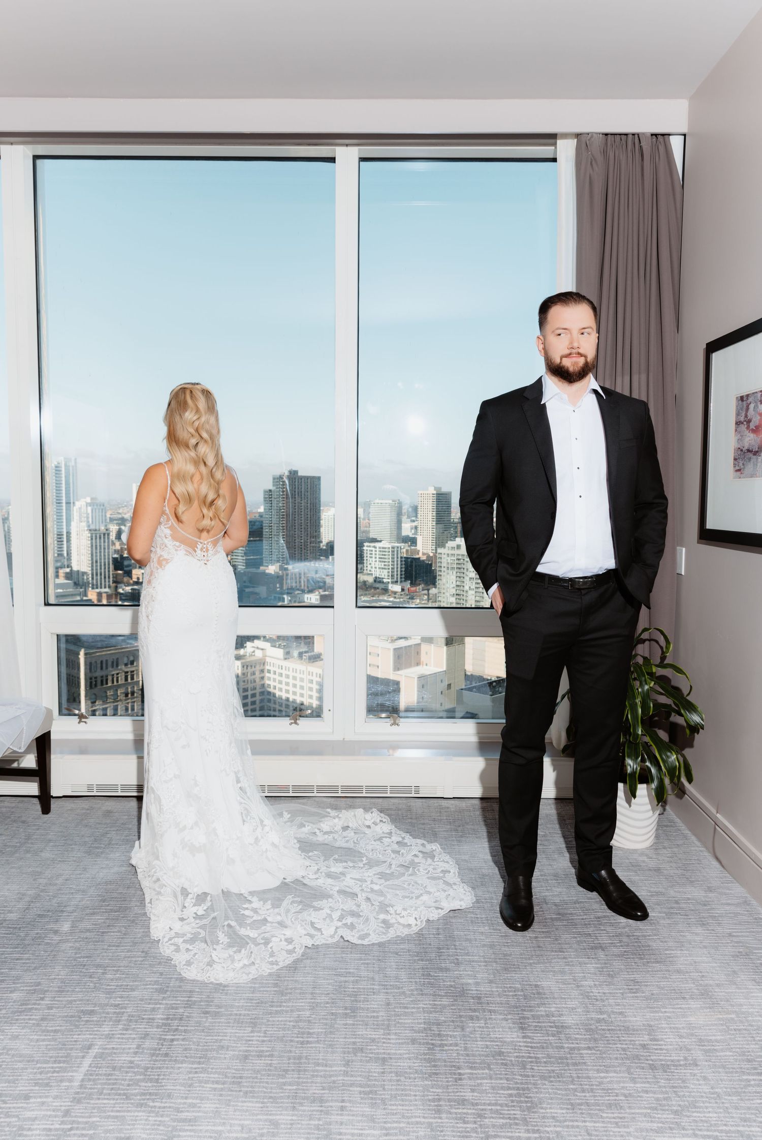 Groom in black tuxedo standing confidently while bride in lace cathedral train gown faces city window.