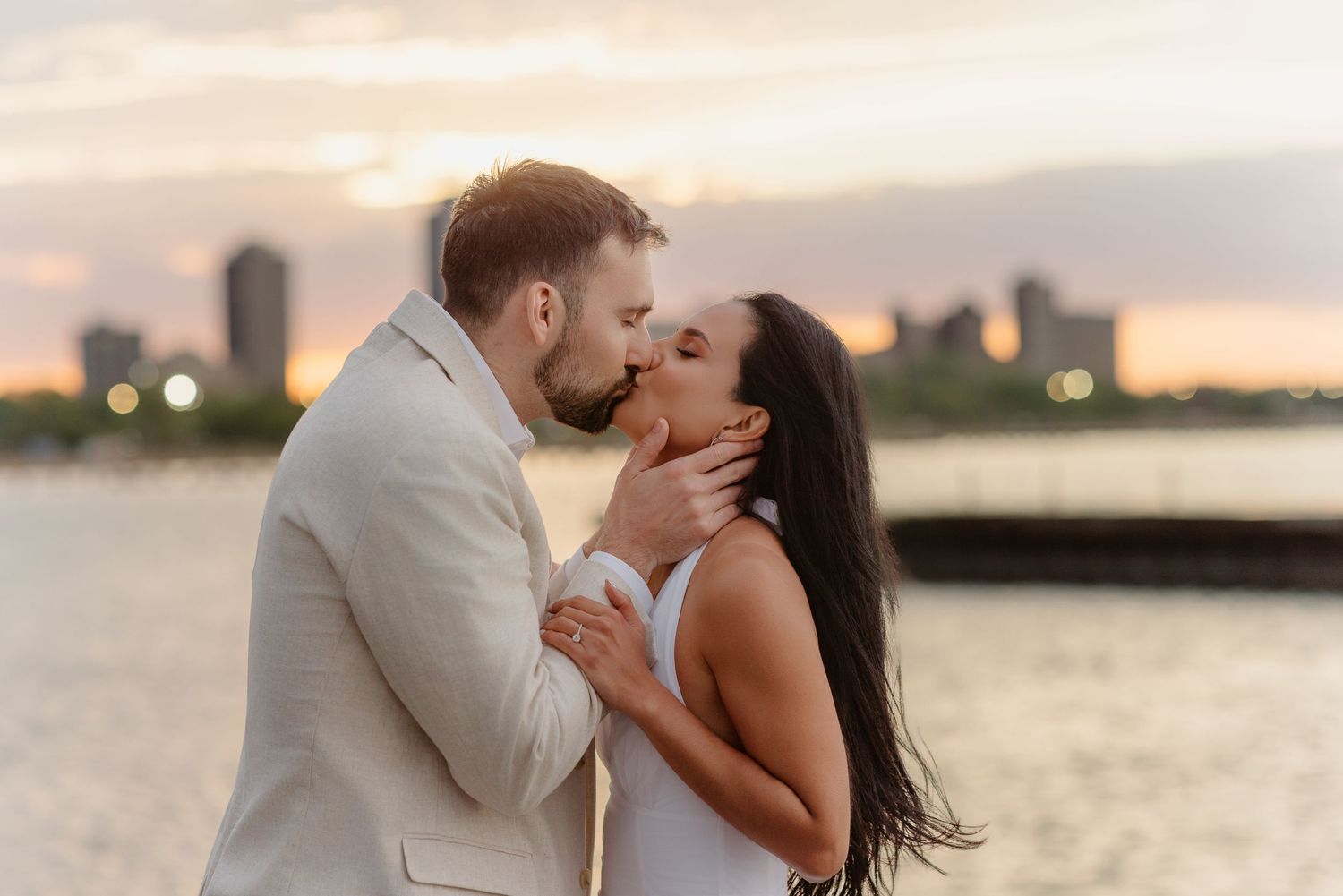 Engaged couple kissing tenderly at golden hour with urban skyline reflected on water.