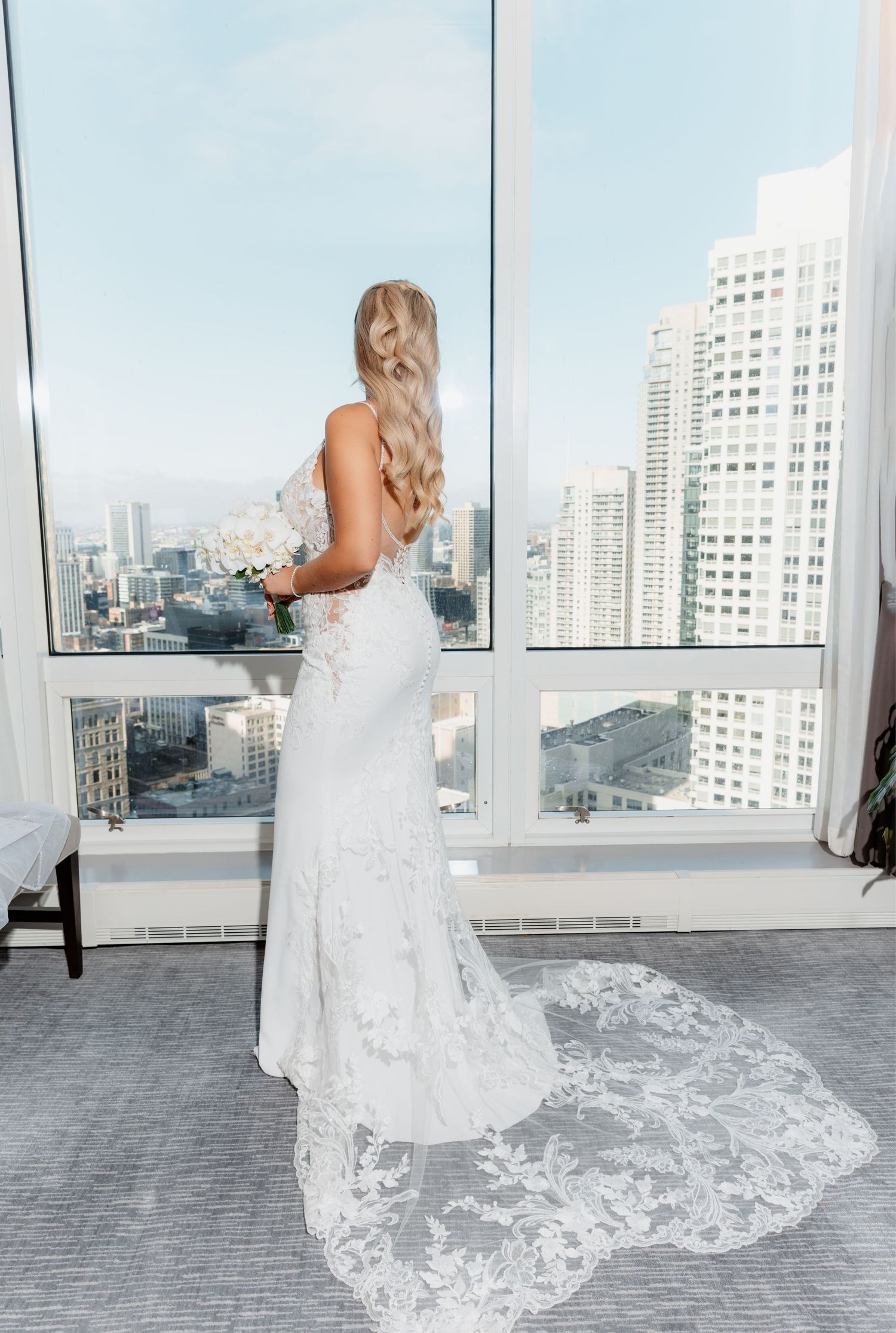 Bride in fitted lace gown with cathedral train views city skyline through large panoramic hotel windows.