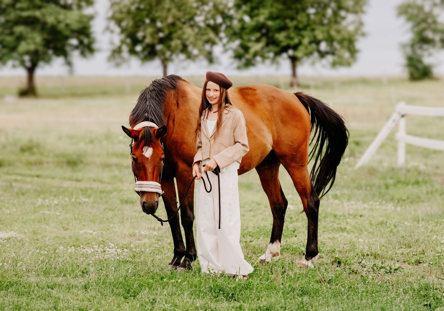 Woman in beige coat and red beret poses with a grazing chestnut horse in a green field.