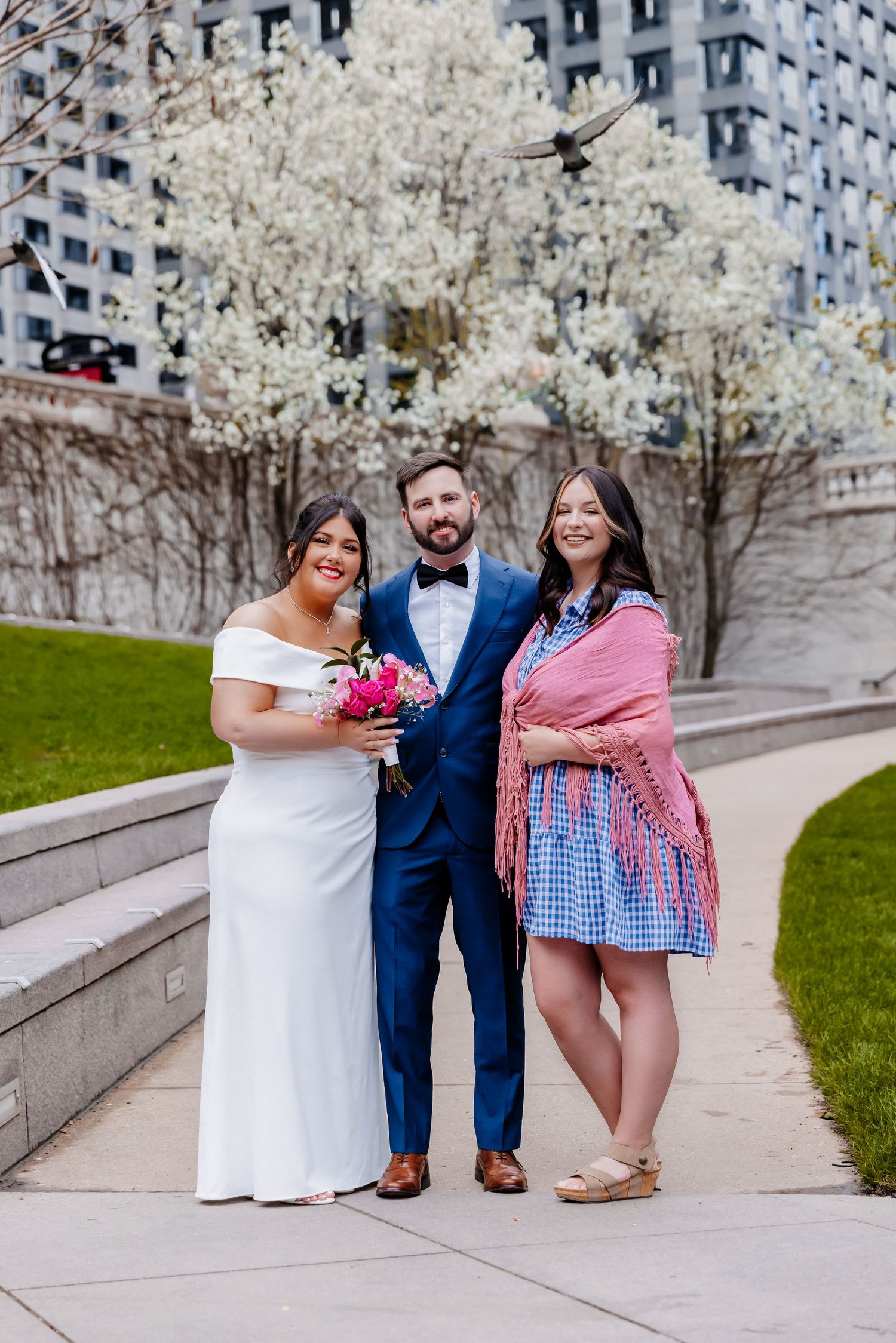 Bride in white gown and groom in blue suit pose with smiling guest near spring blooming trees.