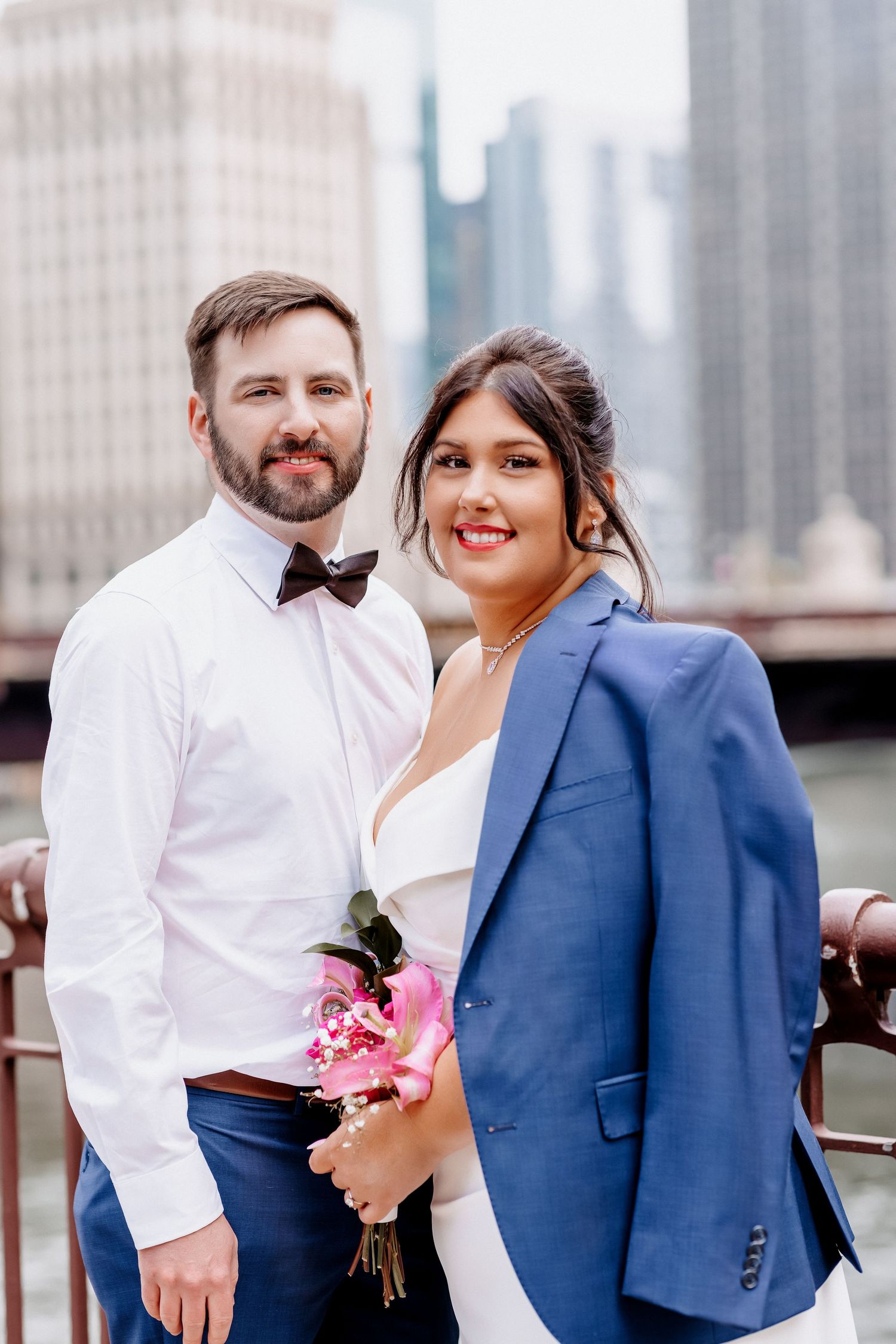 Couple poses on Chicago bridge, groom in navy suit with bow tie, bride holding pink bouquet in blue blazer.