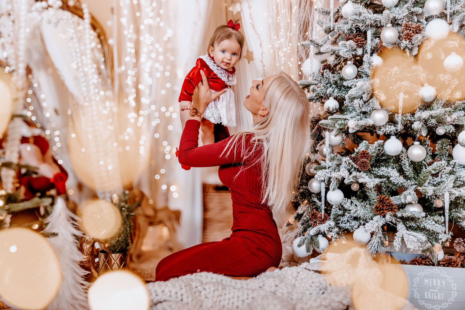 Blonde mother in red outfit lifts smiling toddler near decorated Christmas tree with white ornaments and fairy lights.