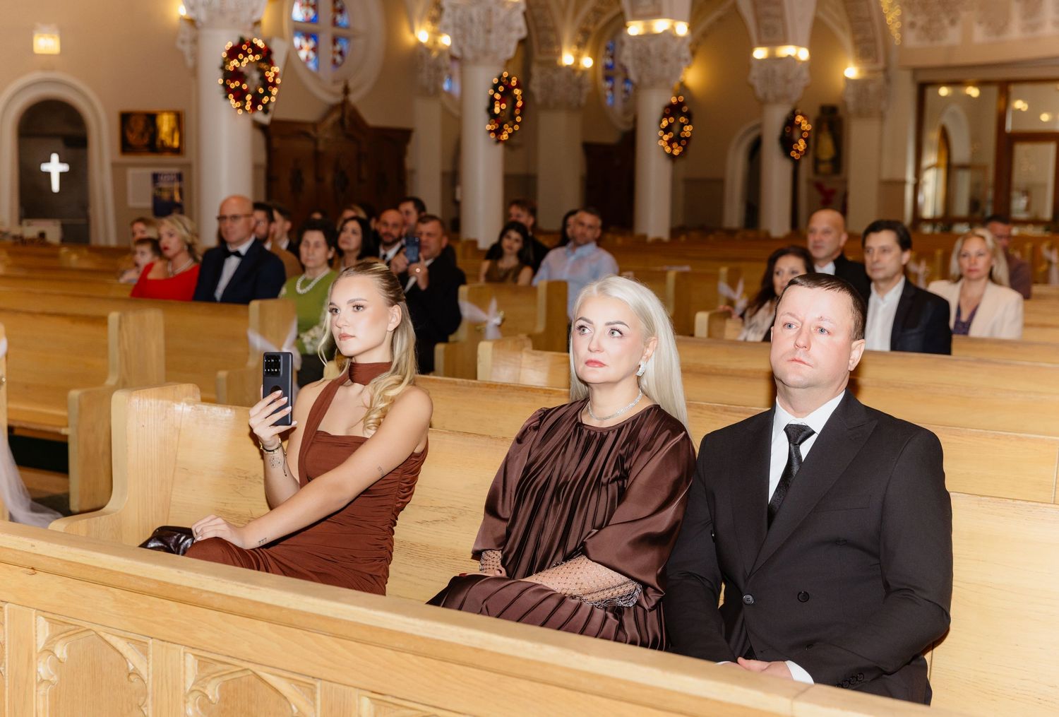 Wedding guests seated in church pews watching ceremony including woman in brown dress in foreground.