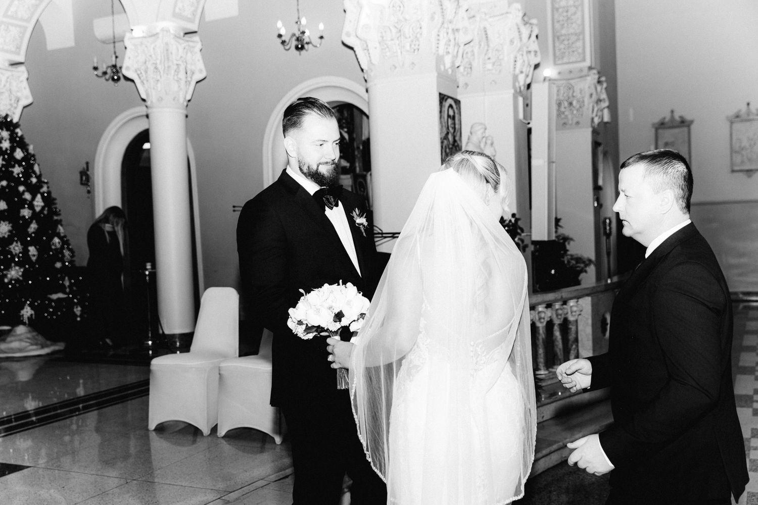 Black and white photo of groom smiling at veiled bride holding white bouquet in decorated church.