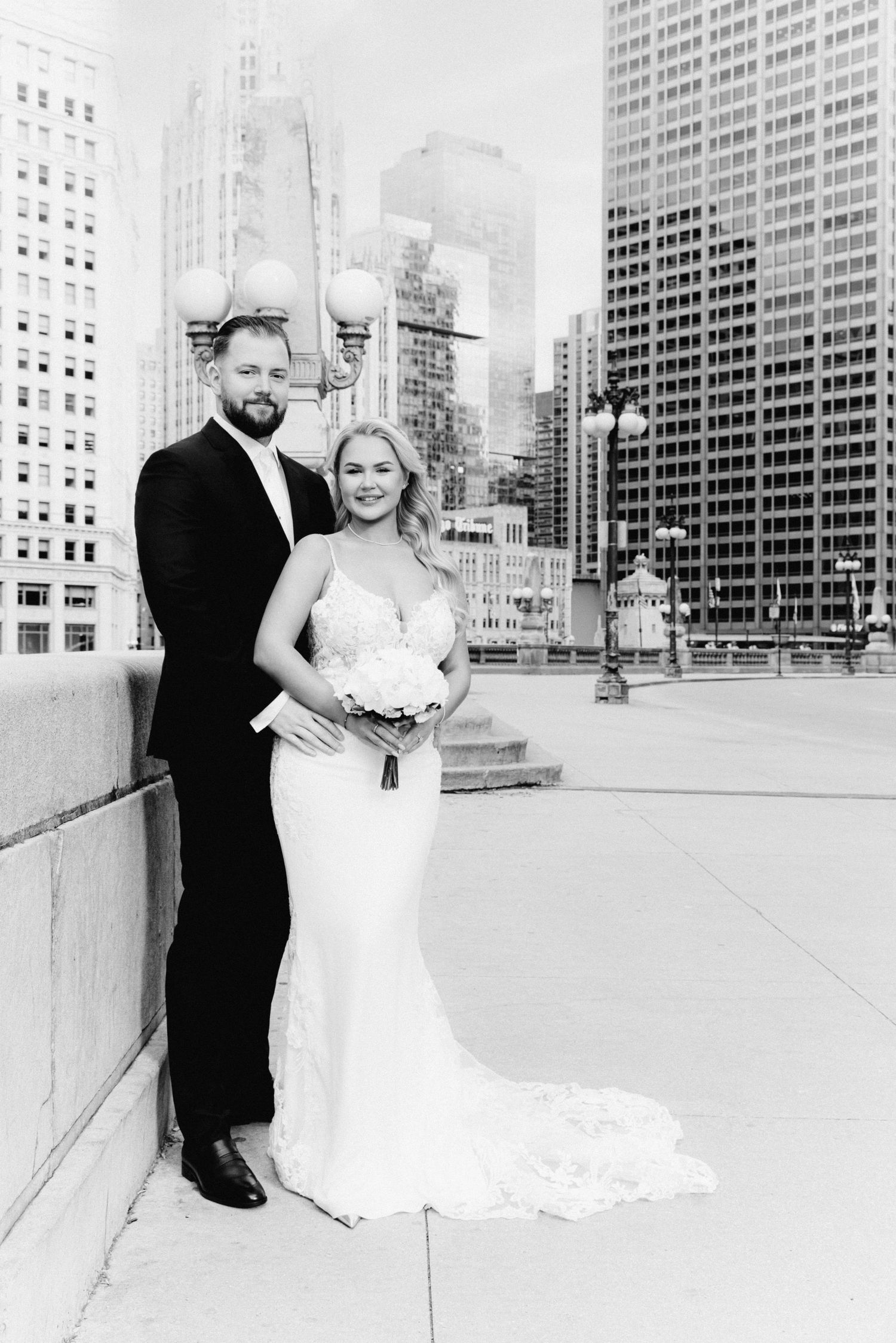 Black and white photo of bride and groom posing on Chicago bridge with city skyline backdrop.