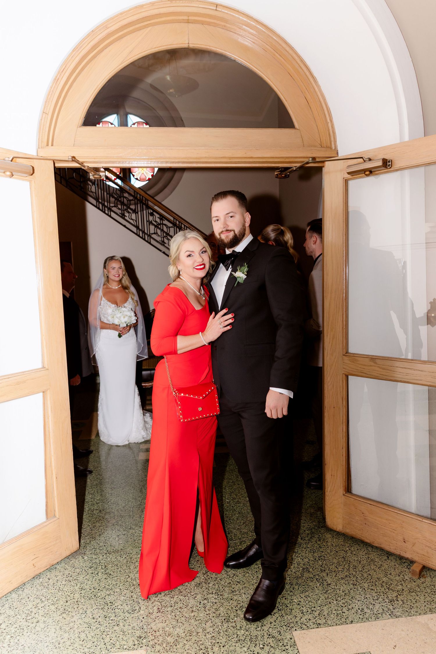 Bearded groom in black tuxedo and woman in red dress stand in arched church doorway with bride visible behind.