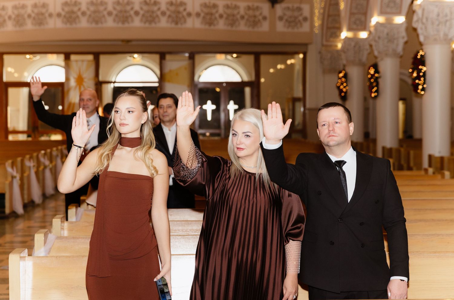 Three elegantly dressed wedding guests wave while standing in a beautifully decorated church aisle.