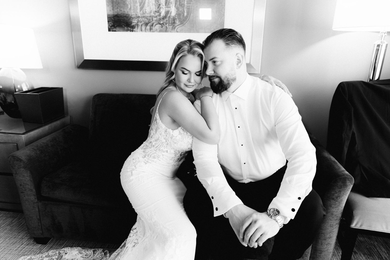 Black and white photo of bride in lace dress leaning on groom while seated on armchair.