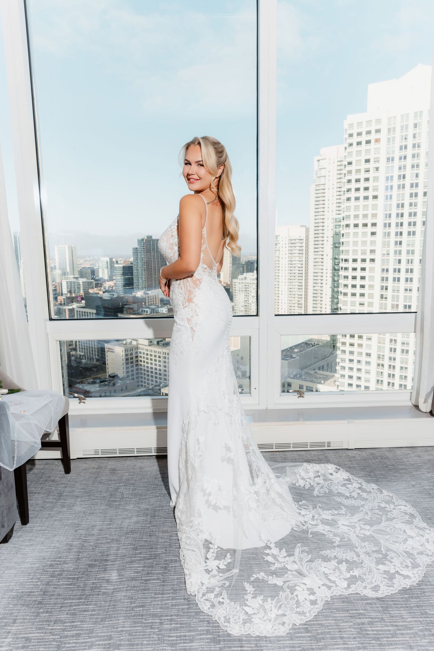 Bride in backless lace wedding gown with train smiling over shoulder before floor-to-ceiling city view windows.