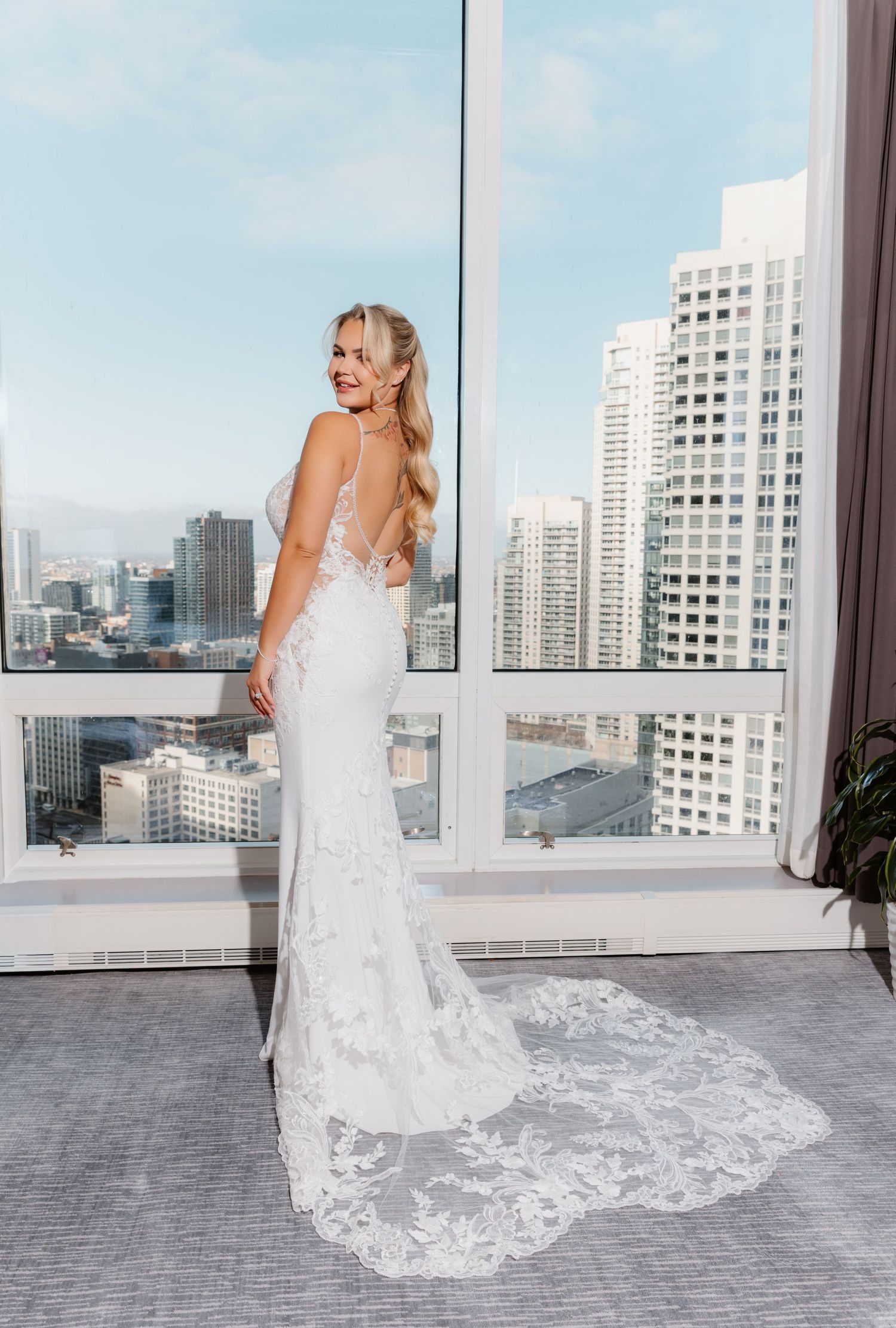 Bride in fitted lace wedding gown with long train standing by window with urban skyline view.