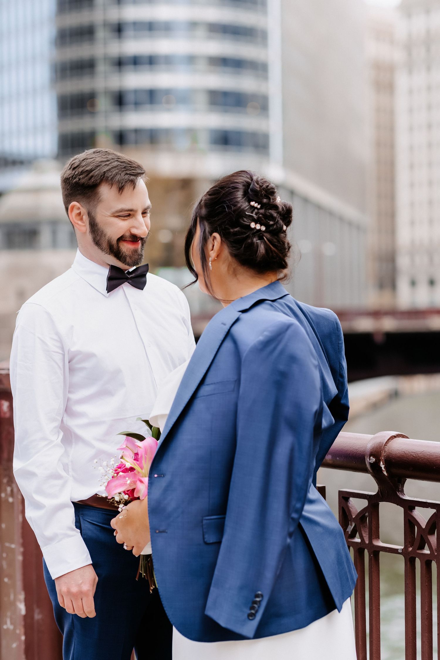 Groom in white shirt and bow tie smiles at bride in blue blazer on Chicago bridge with city skyline.