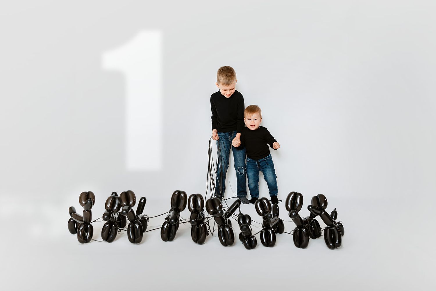 Two boys in black tops with black balloon animal dogs beside a number 1 on white background.