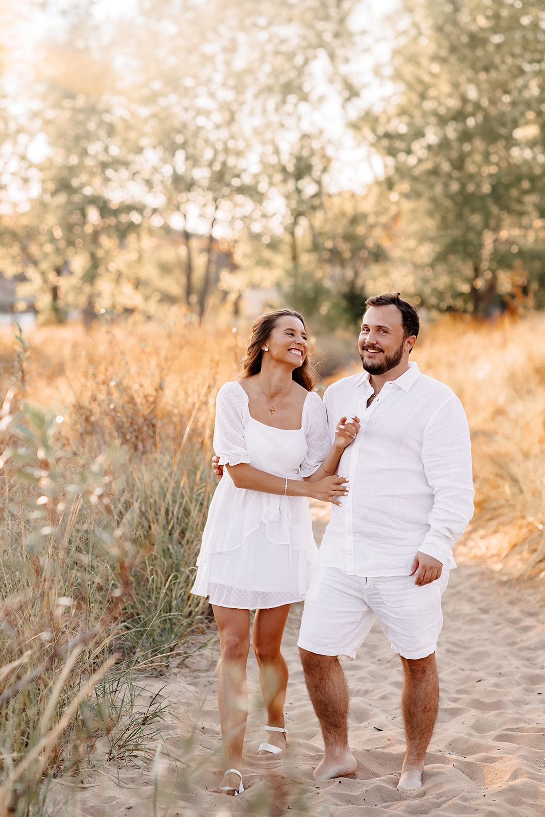 Happy couple in matching white outfits walking on sandy beach path surrounded by golden grass at sunset.