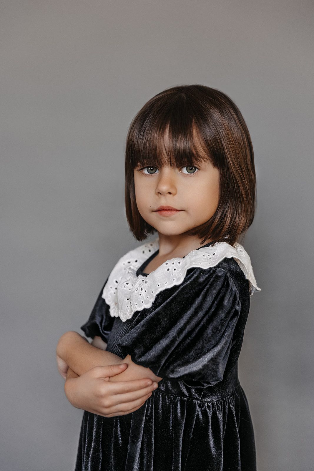 Young girl in dark velvet dress with white lace collar stands arms crossed on gray background.