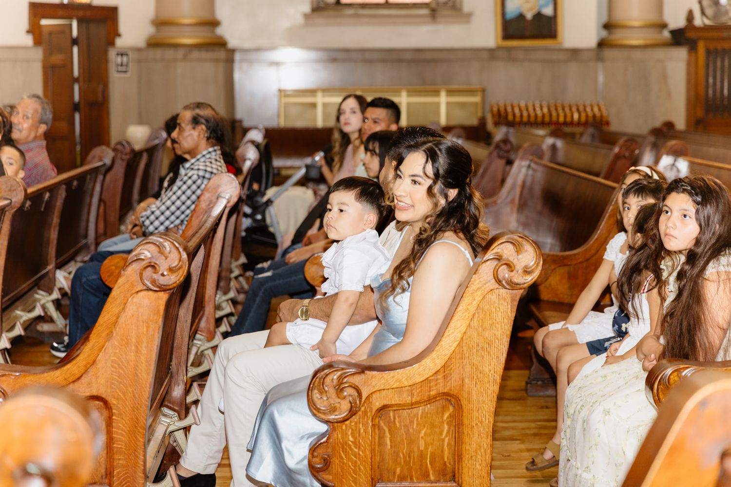 Families seated in wooden church pews during a religious ceremony, with children dressed in white formal attire.