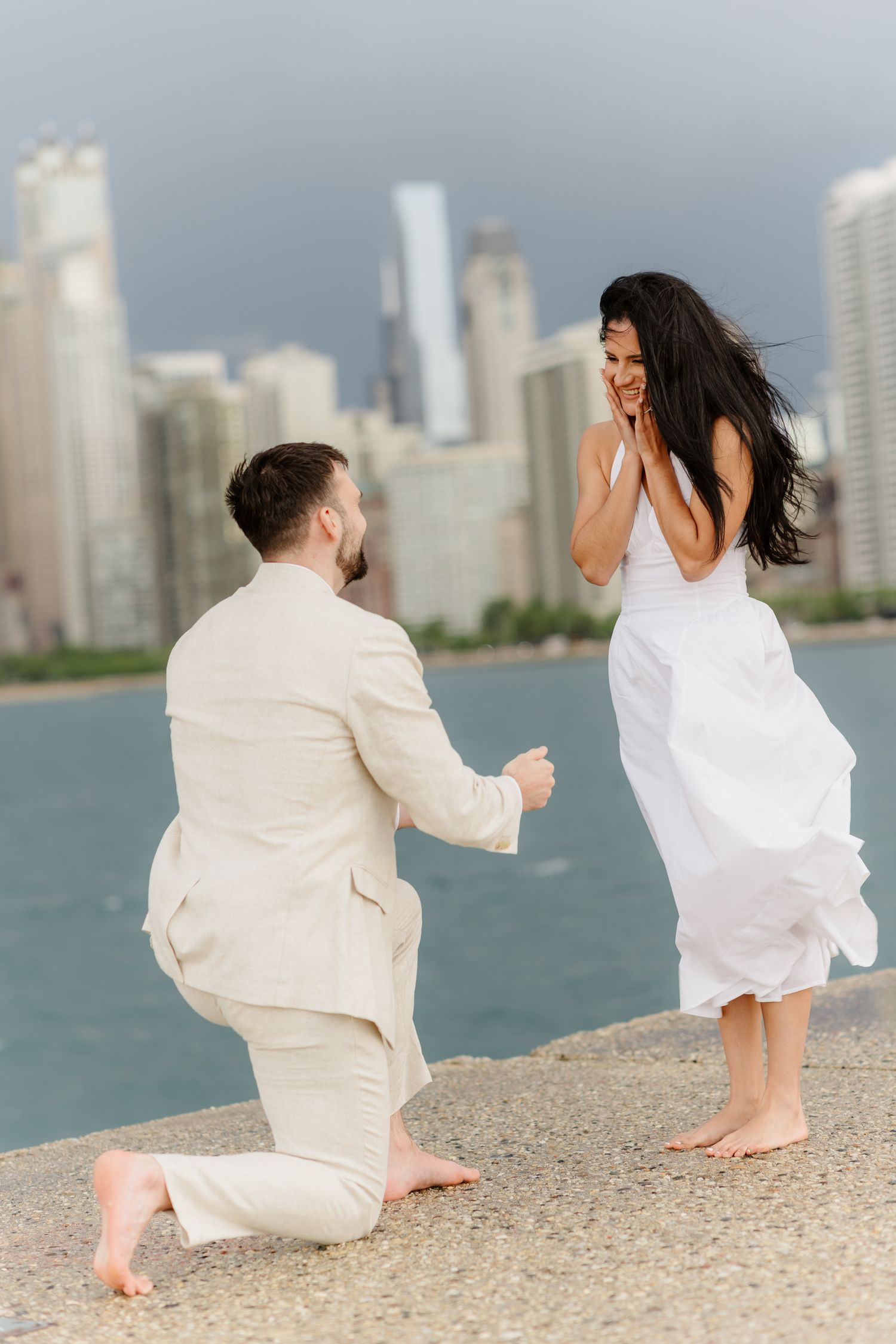 Man proposes on one knee to woman in white dress by waterfront with city skyline backdrop.