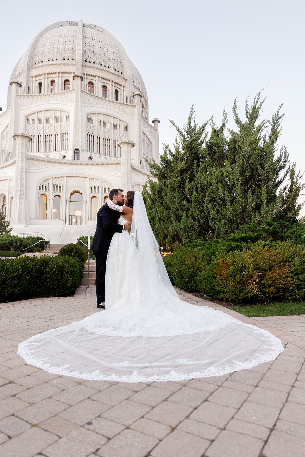 Bride and groom kiss on stone plaza with the majestic Bahai Temple dome and lace cathedral veil on display.