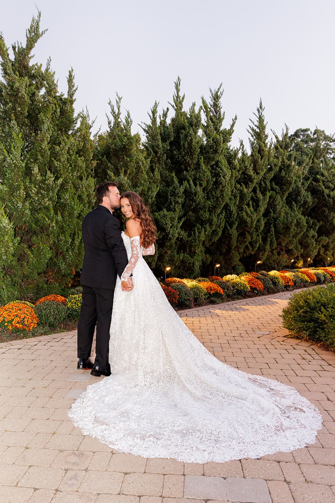 A bride in a lace gown with a long train and groom in a tuxedo share a romantic moment in a garden.