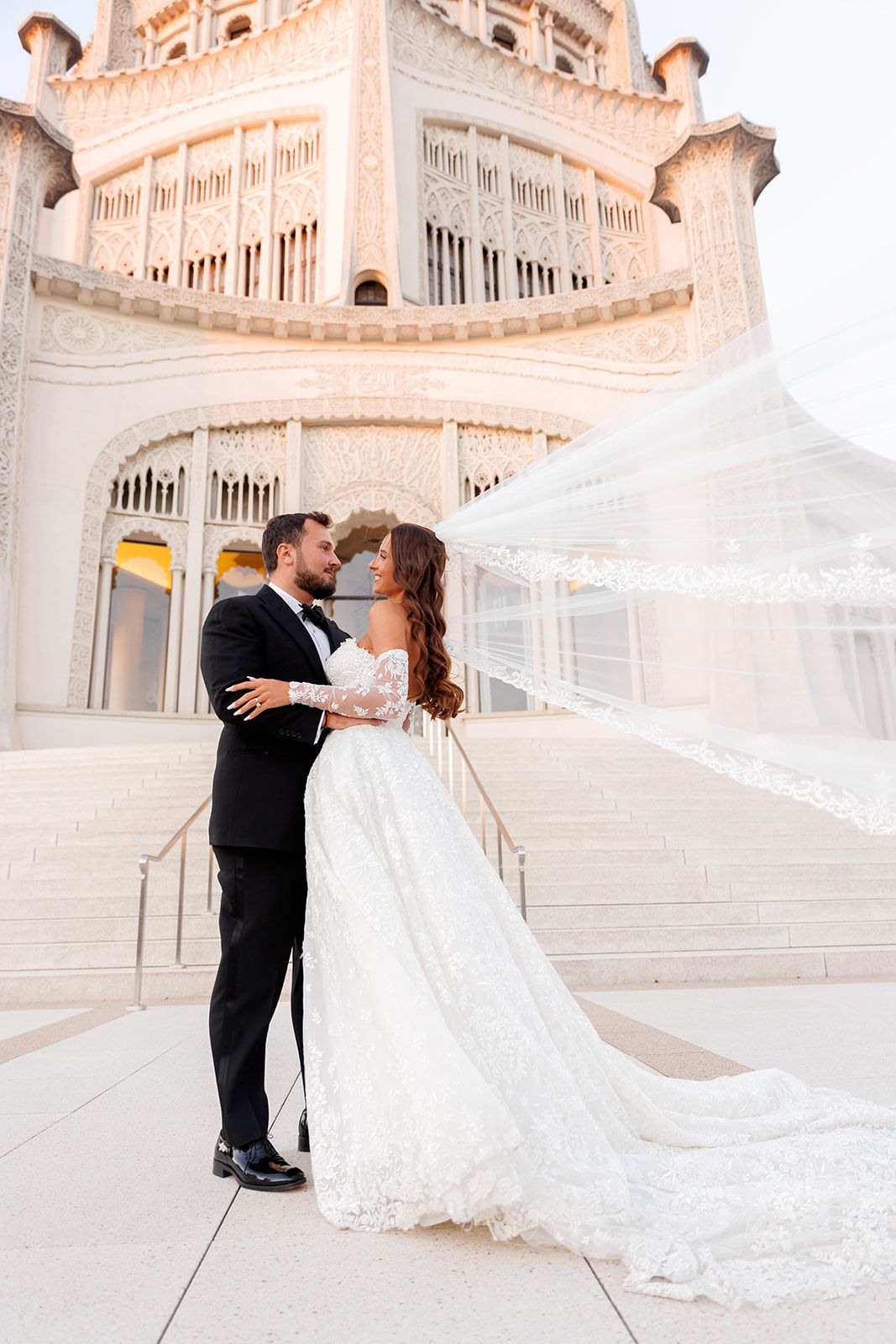 Bride and groom embrace in front of ornate white Bahai Temple, her lace veil flowing dramatically in the wind.