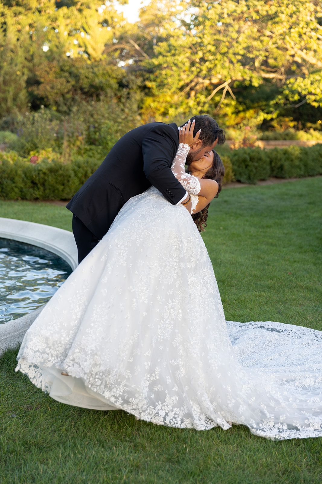 Groom dips bride in a romantic kiss on garden lawn, bride wearing floral lace ball gown near a fountain.