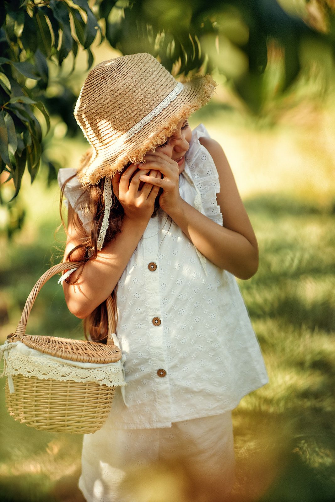 A young girl in a white linen outfit and straw hat holds a wicker basket while standing in a sunlit orchard.