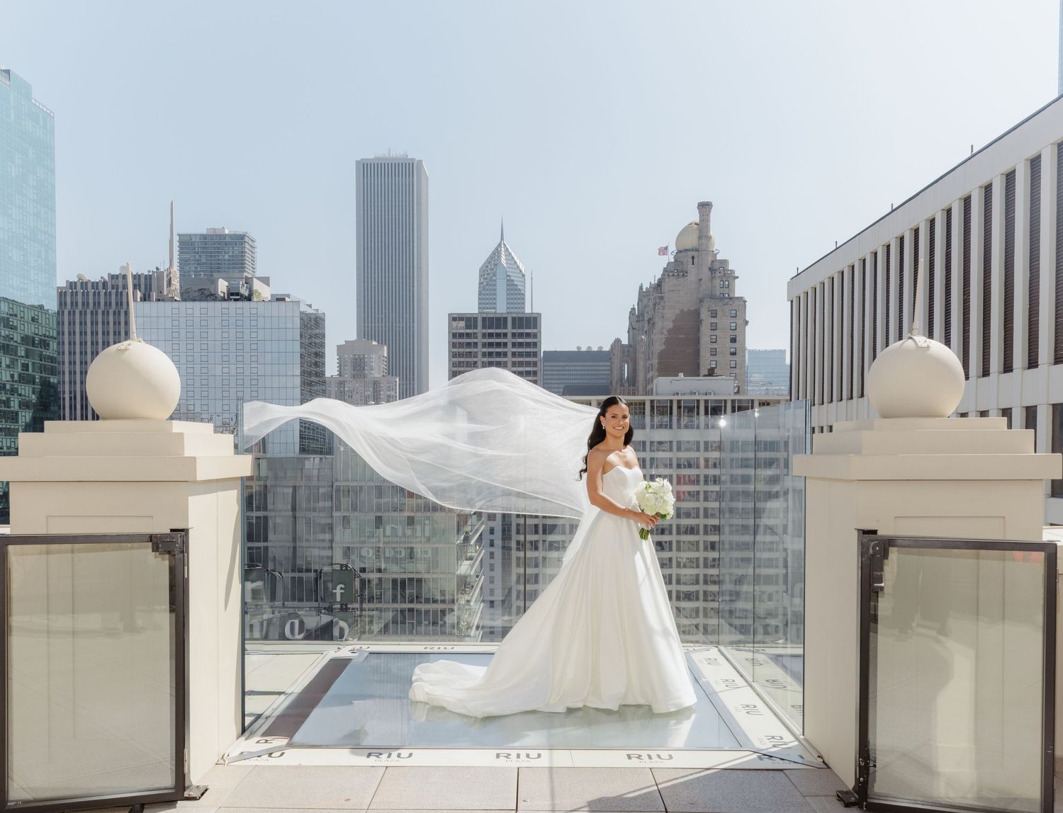 Bride in flowing white gown with long veil billowing on rooftop overlooking Chicago city skyline on sunny day.