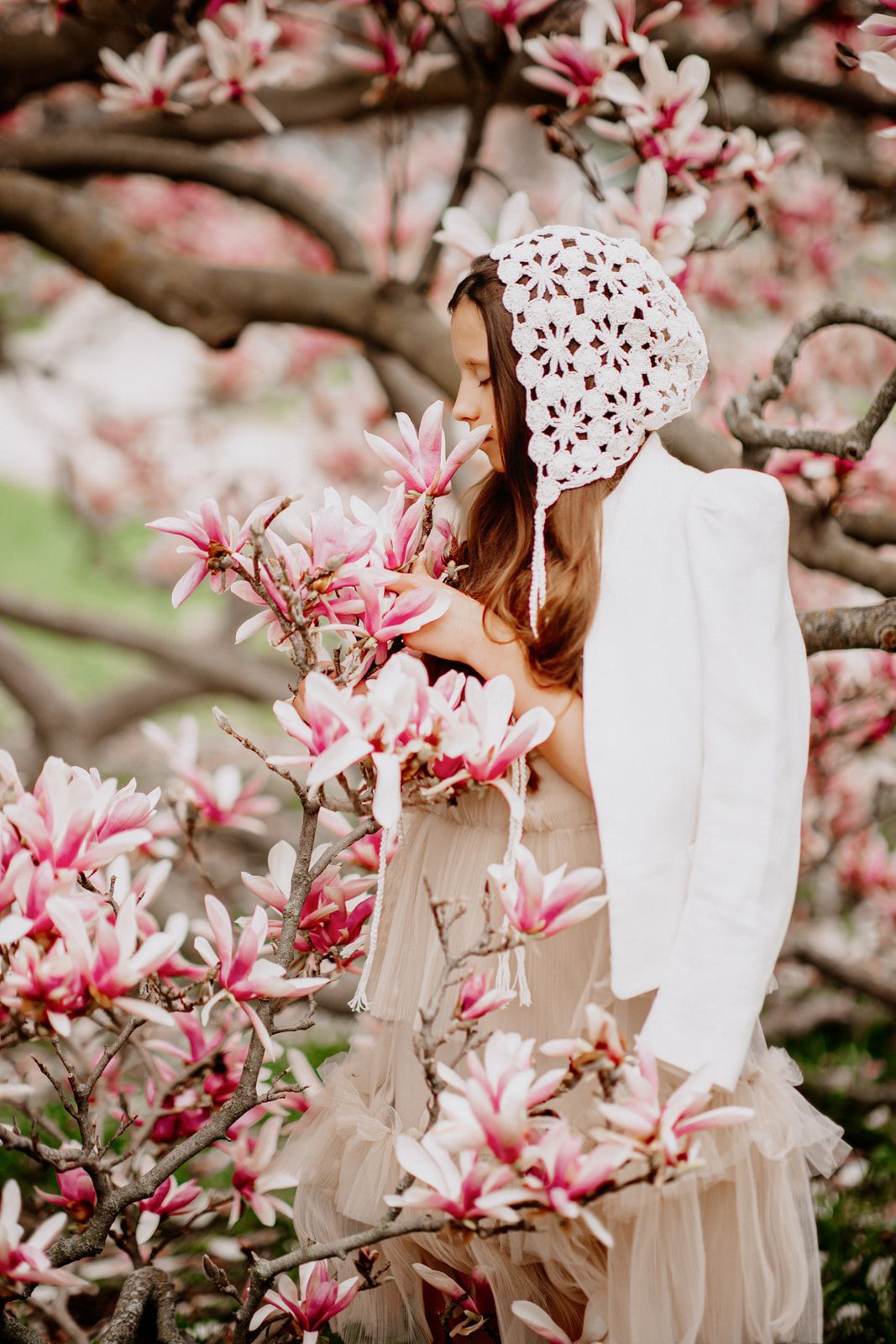Young woman in white crochet bonnet and dress admiring pink magnolia blossoms on a flowering tree in spring.