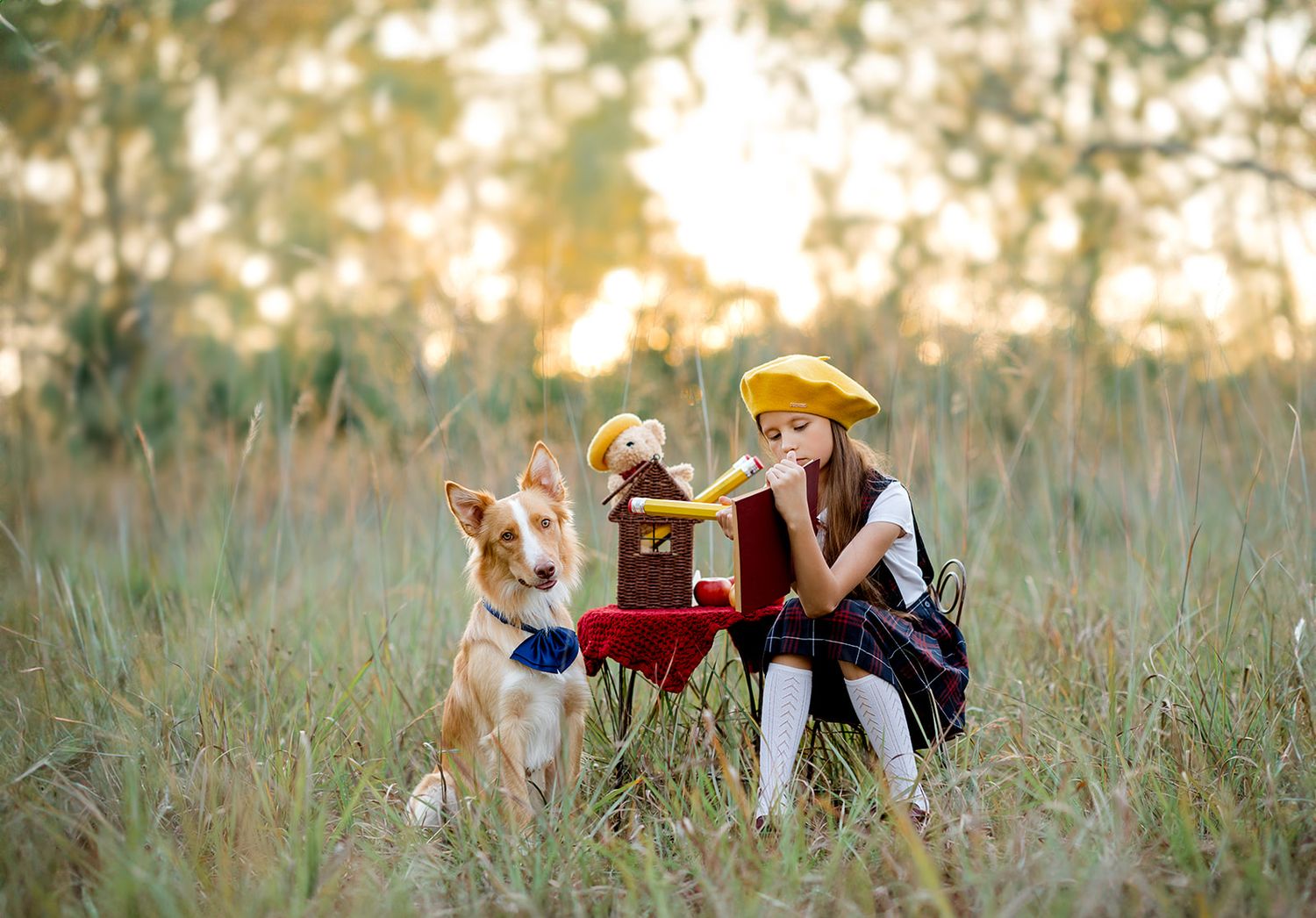 Young girl in yellow beret sits in field with golden retriever dog at small red table at sunset.