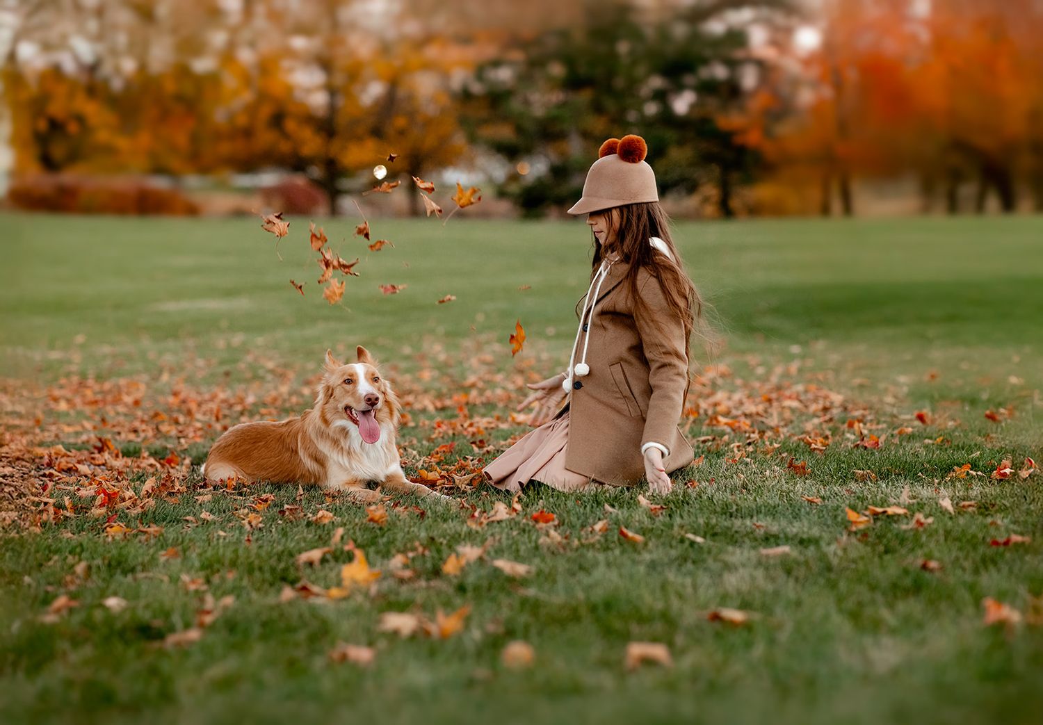 Woman in beige coat and hat plays with Corgi dog among fallen autumn leaves in a lush green park.