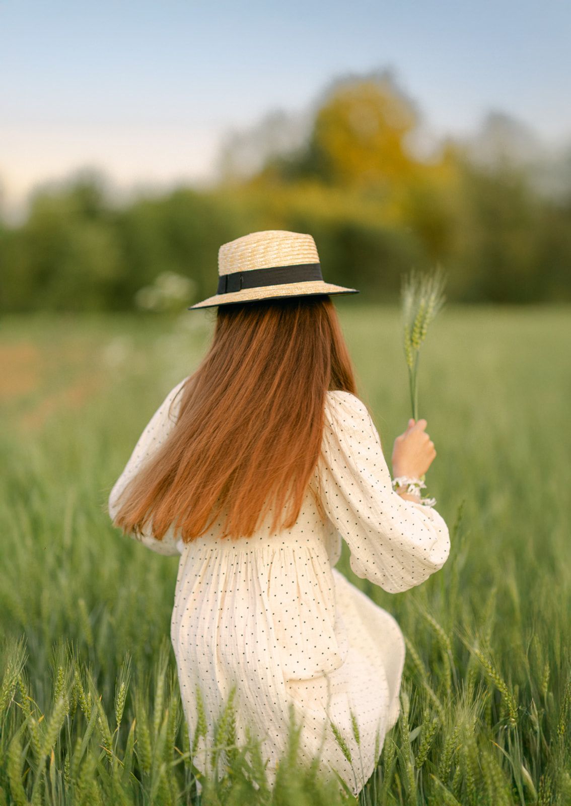 A young woman with long red hair wearing a straw hat and white dress walks through a lush green wheat field.