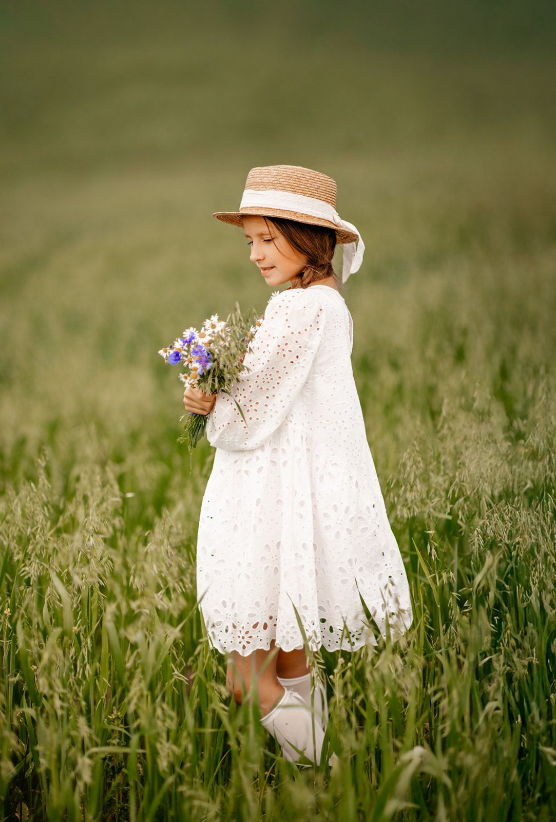 Smiling girl in white eyelet dress and straw hat holding colorful wildflowers in a green meadow.