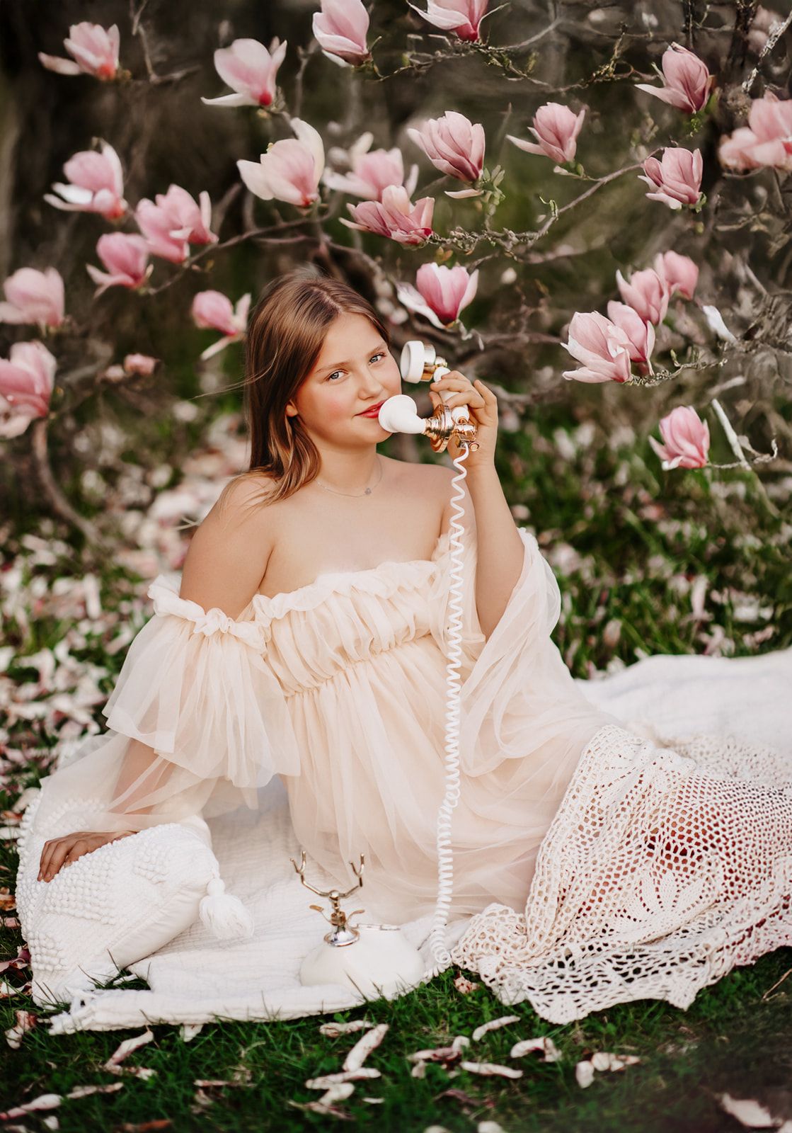 Woman in off-shoulder white dress sitting among pink magnolia blooms holding vintage phone.