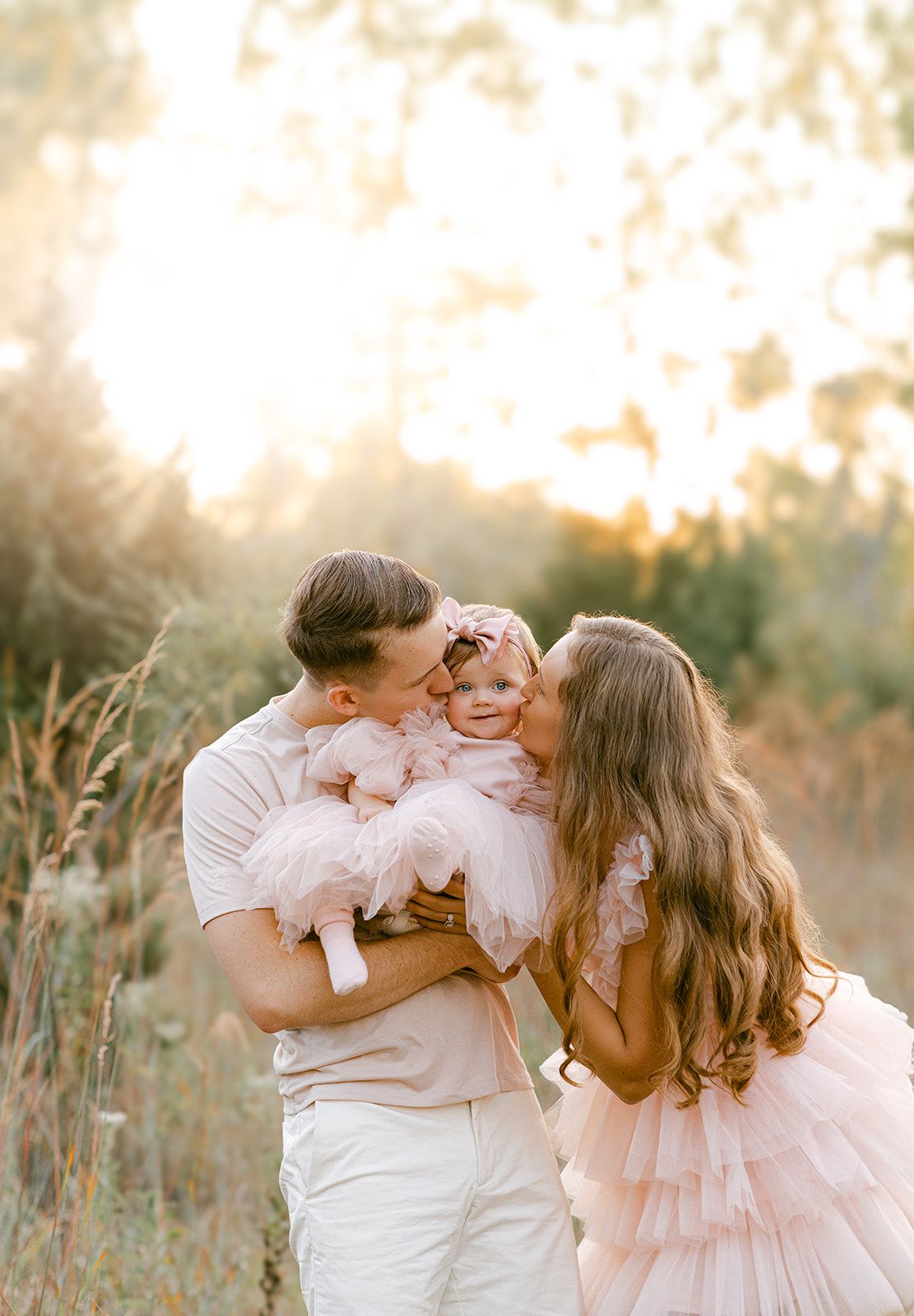 A couple kisses their baby girl in a pink tutu dress during a golden hour outdoor photo session.