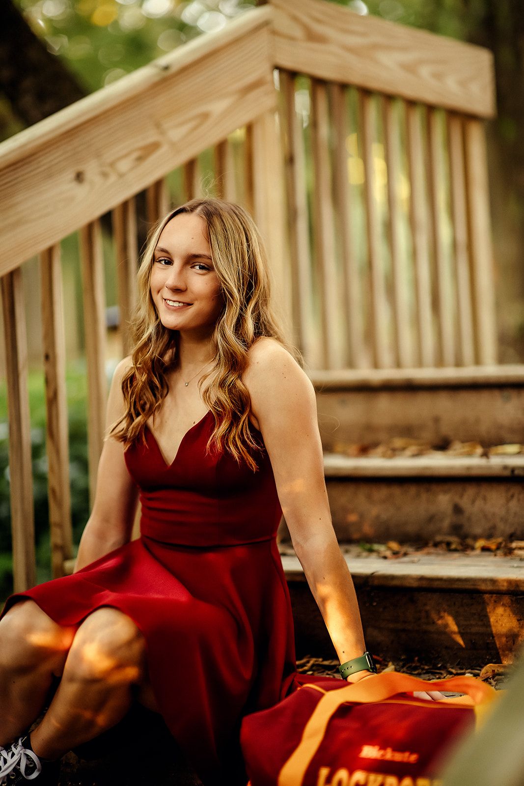 Smiling young woman in red dress sits on wooden outdoor stairs surrounded by autumn leaves.