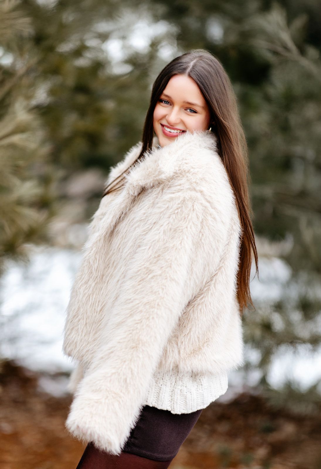 Cheerful young woman in white faux fur coat smiling over shoulder in snowy evergreen forest.