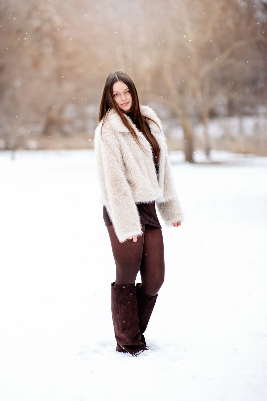 Young woman in white fur coat, dark pants, and boots standing in snowy winter park landscape.