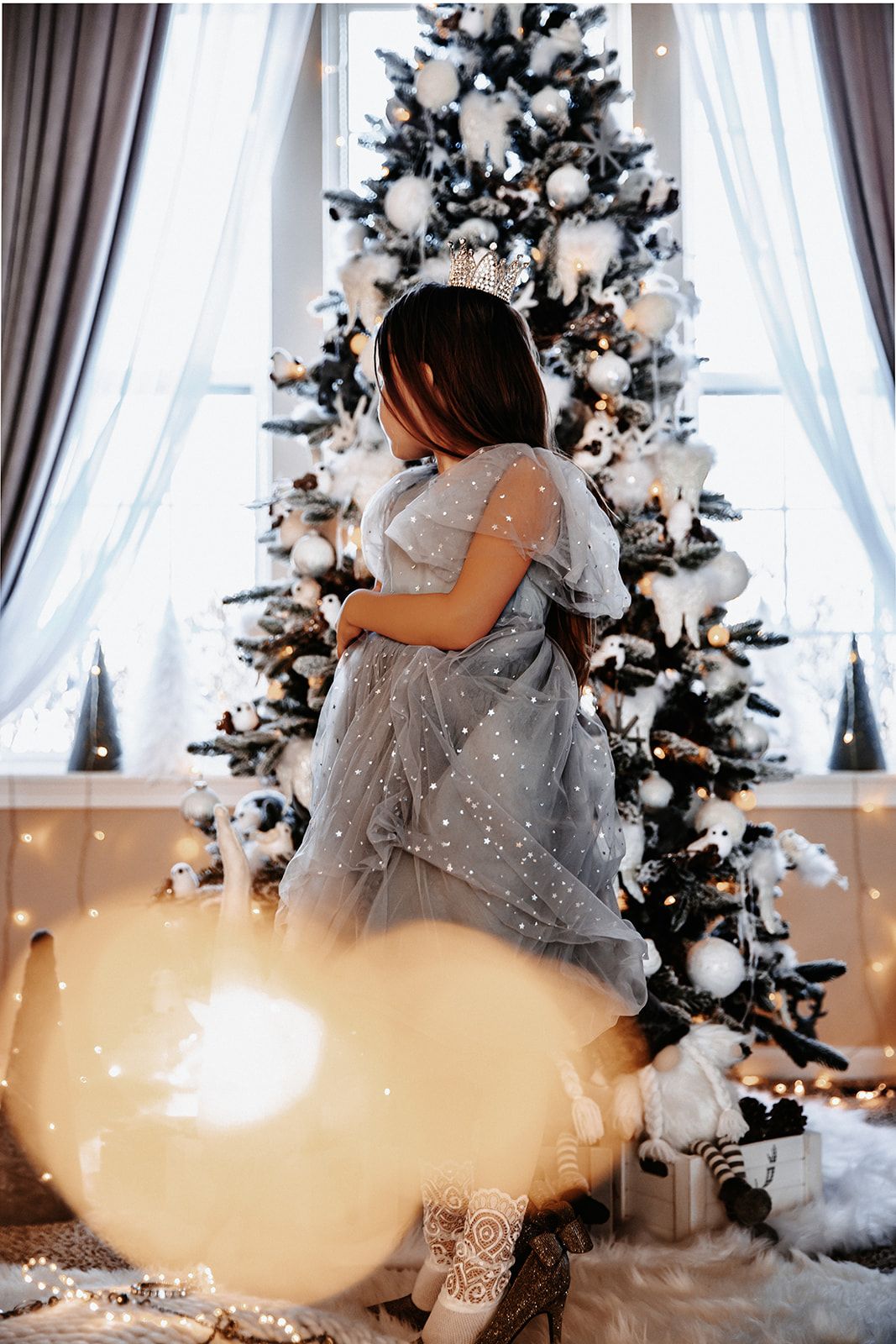 A young girl in a silver tulle gown and crown poses before a decorated Christmas tree with white ornaments.