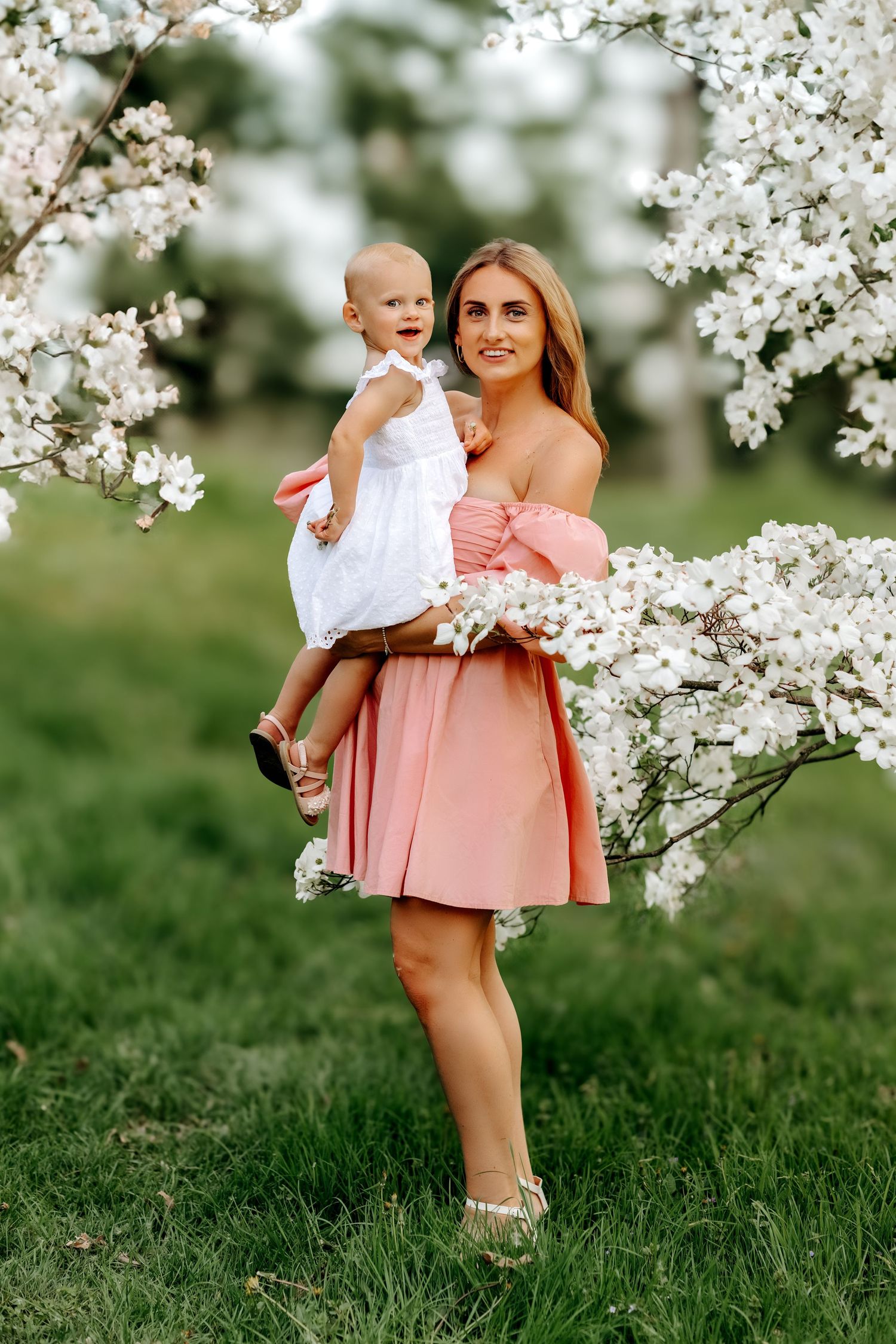 Elegant woman in pink dress holding laughing baby near white flowering spring blossoms outdoors.