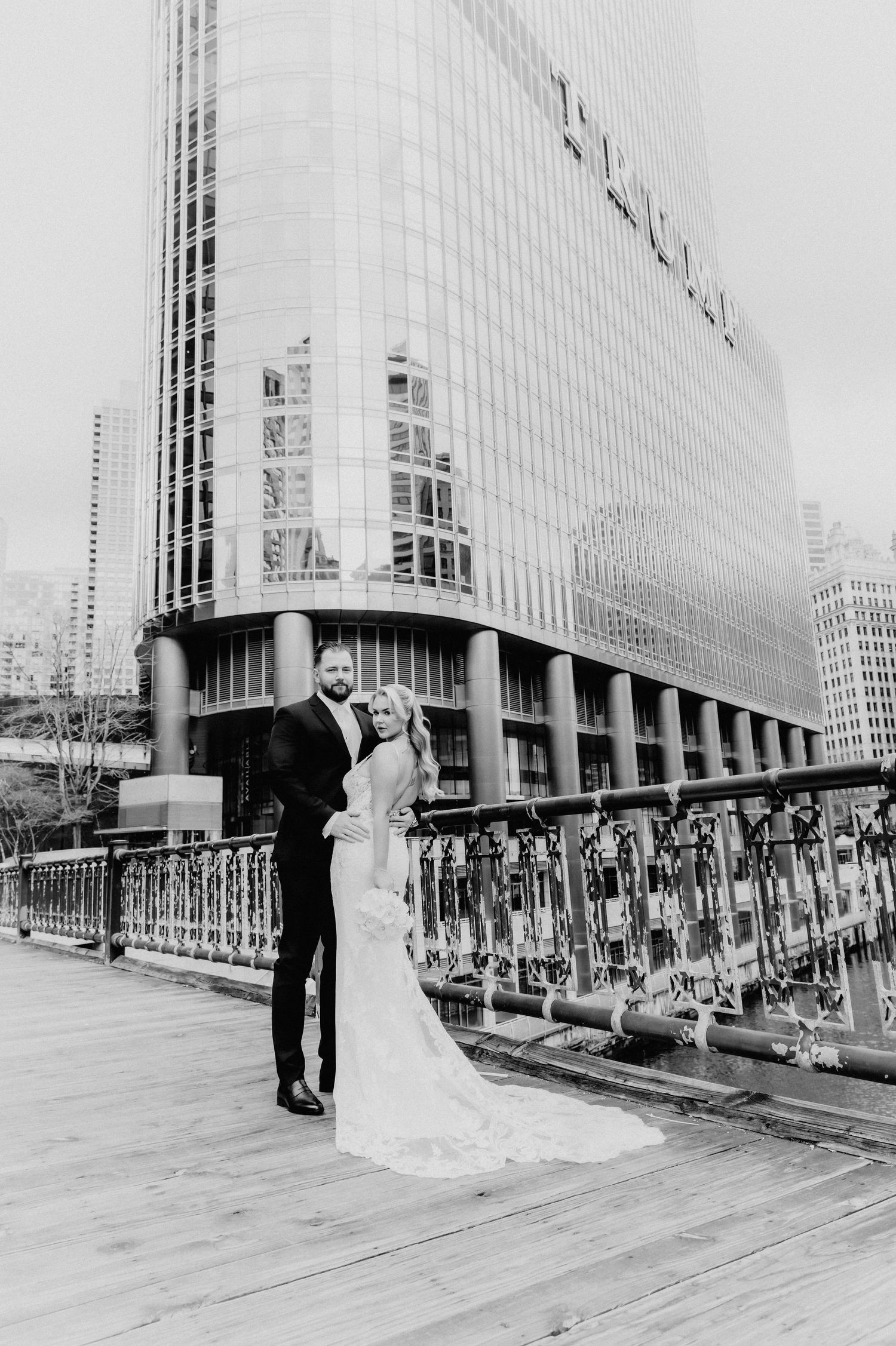 Black and white wedding portrait of couple on Chicago riverwalk with glass skyscraper behind them.