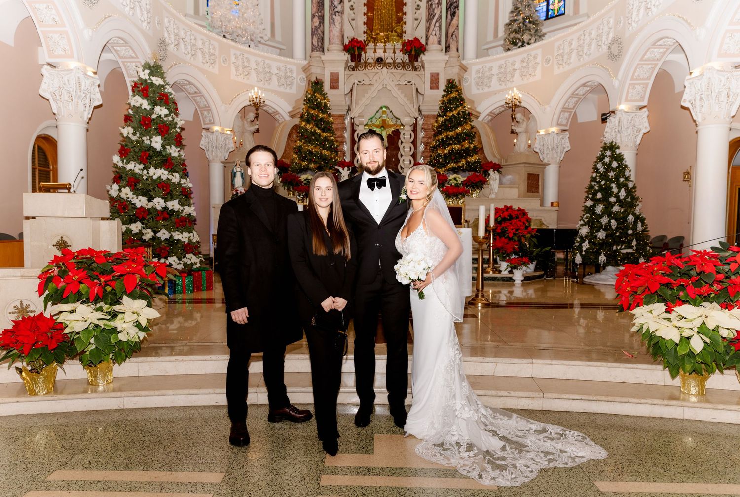 Wedding couple and two guests smile together in a festively decorated church with Christmas trees.