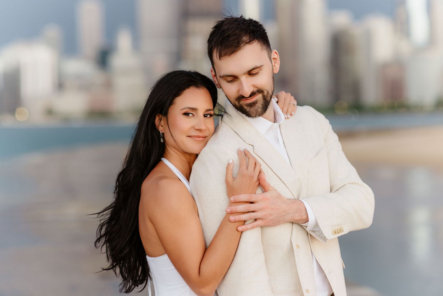 Couple sharing a tender moment on Chicago lakefront, woman in white dress embracing man in beige suit.