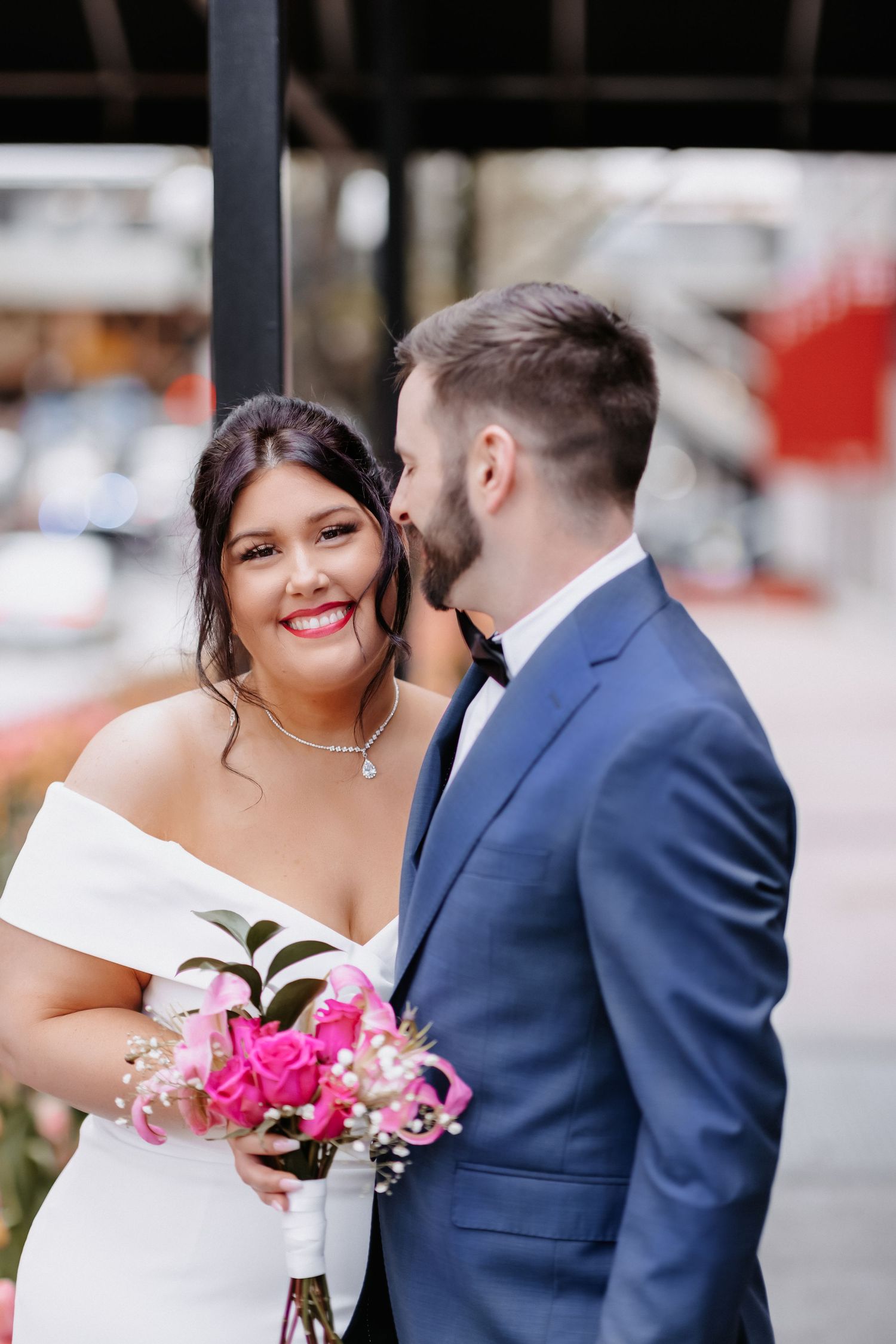 Radiant bride in white dress holds pink roses bouquet while groom in blue suit whispers to her.
