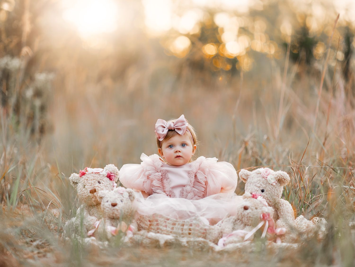Baby girl in pink dress sits in basket with teddy bears in golden field at sunset.